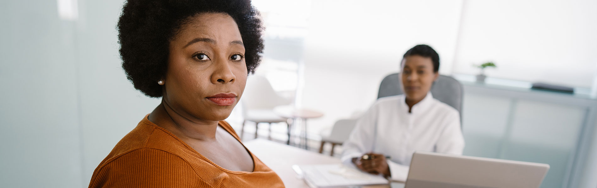 Patient sitting across a desk from her doctor in a bright clinic office, looking directly toward the camera